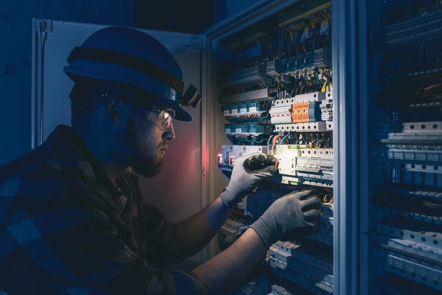 Electrician wearing safety gear working inside an electrical control panel with wiring and circuit breakers.