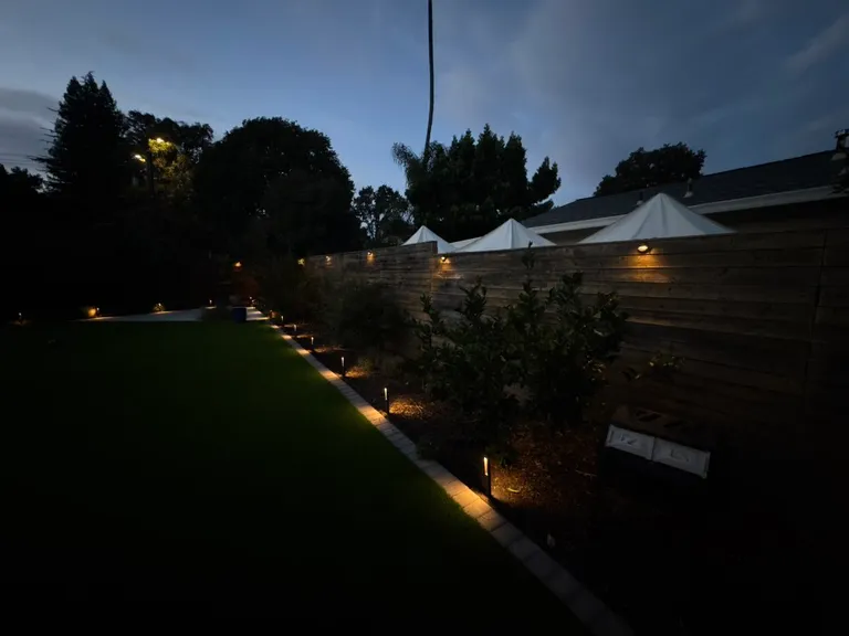 Night view of outdoor garden path lit by small ground and wall lights along a wooden fence and trees.