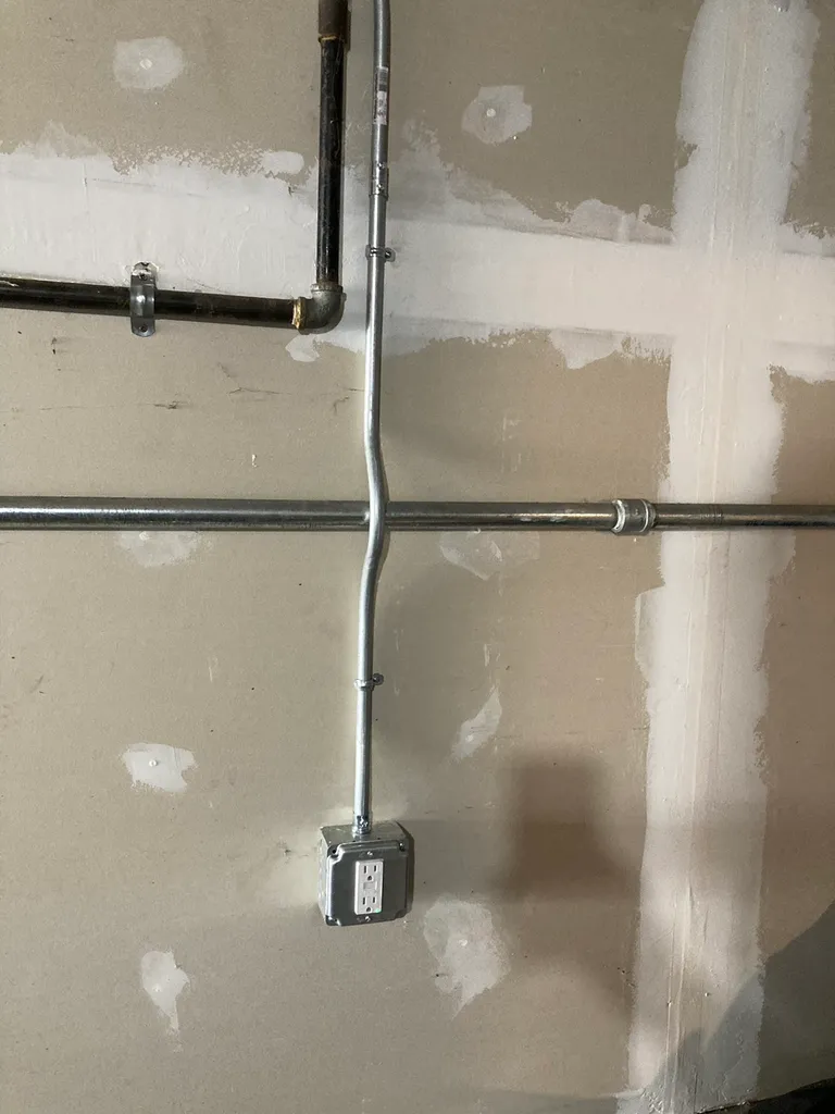Exposed wall with electrical conduit pipes and a metal outlet box with two sockets installed.