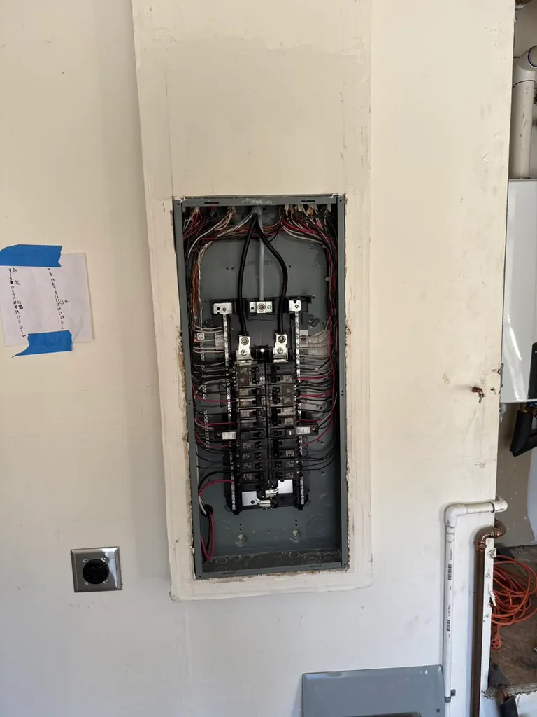 Open electrical panel with circuit breakers and wiring inside a white wall.