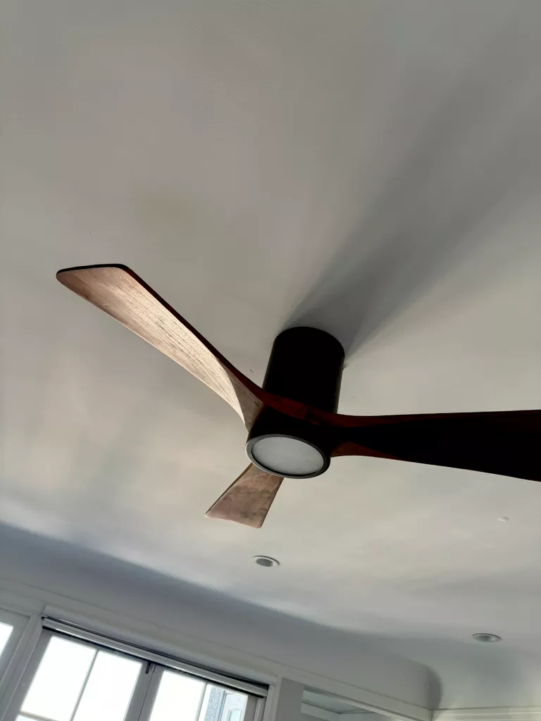 Modern ceiling fan with three wooden blades and a central light fixture against a white ceiling.