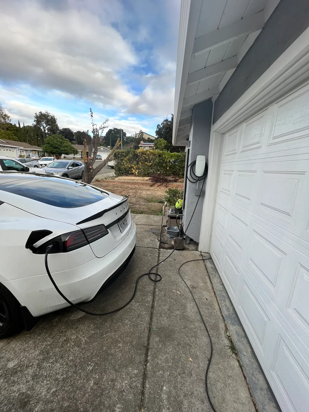 White Tesla electric car charging on a driveway connected to a wall-mounted charger on a gray house.