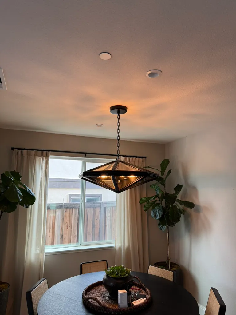 Modern dining room with dark round table, hanging geometric light fixture, plants, and window with curtains.