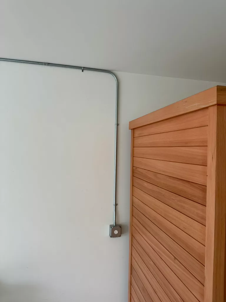 Close-up of a metal electrical conduit and outlet on a plain white wall next to a wooden cabinet.