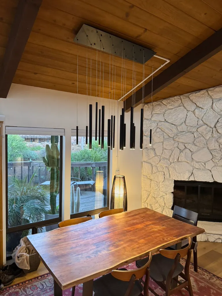Dining area with wooden table, modern pendant lights, stone fireplace, and large windows showing outdoor plants.