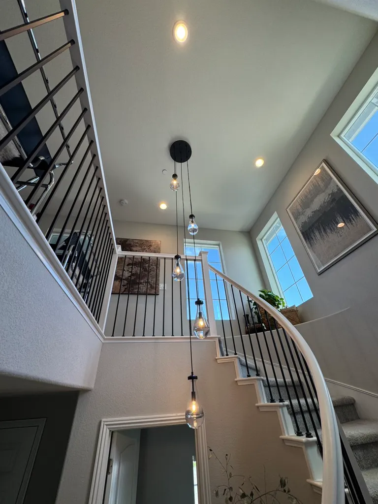 Curved staircase with black metal railing under hanging light bulbs in a bright modern home.