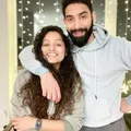 A man and woman smiling, the man hugs the woman from behind with a gray hoodie on.