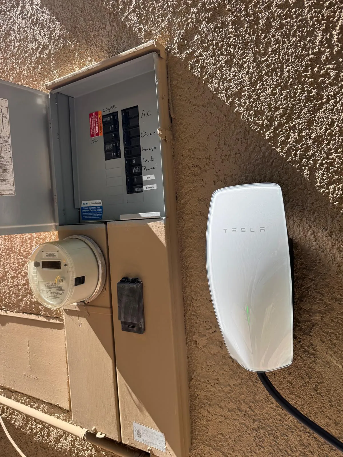 Open electrical panel next to a Tesla electric vehicle charger mounted on a stucco wall.