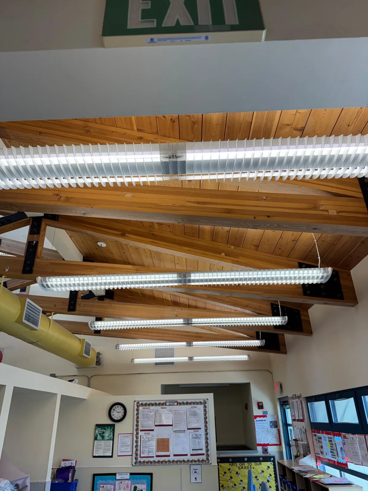 Classroom ceiling with wooden beams, fluorescent lights, exit sign, and a bulletin board on a wall.
