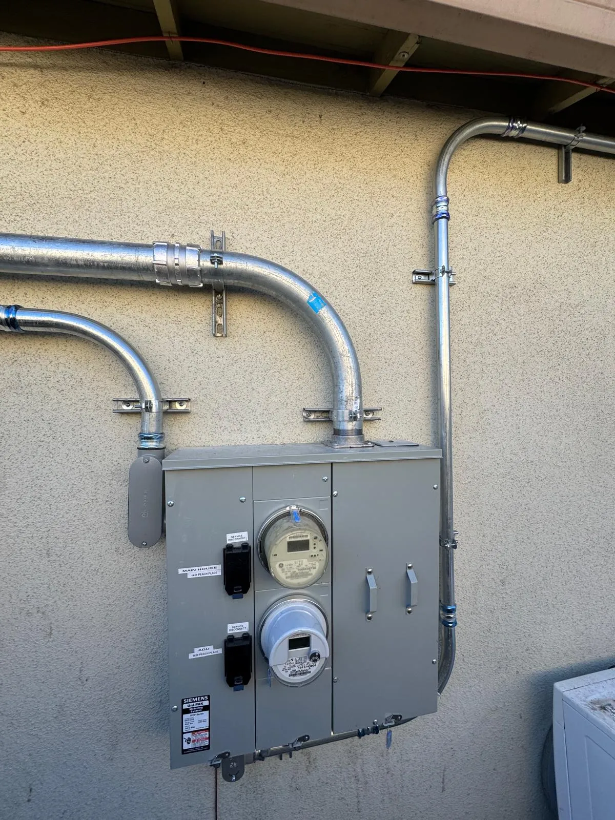 Gray electrical meter panel with two meters mounted on a beige exterior wall with metal conduit pipes.