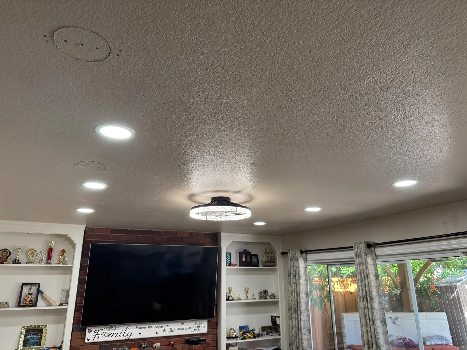 Living room ceiling with recessed and central lighting above a large TV and shelves with trophies.
