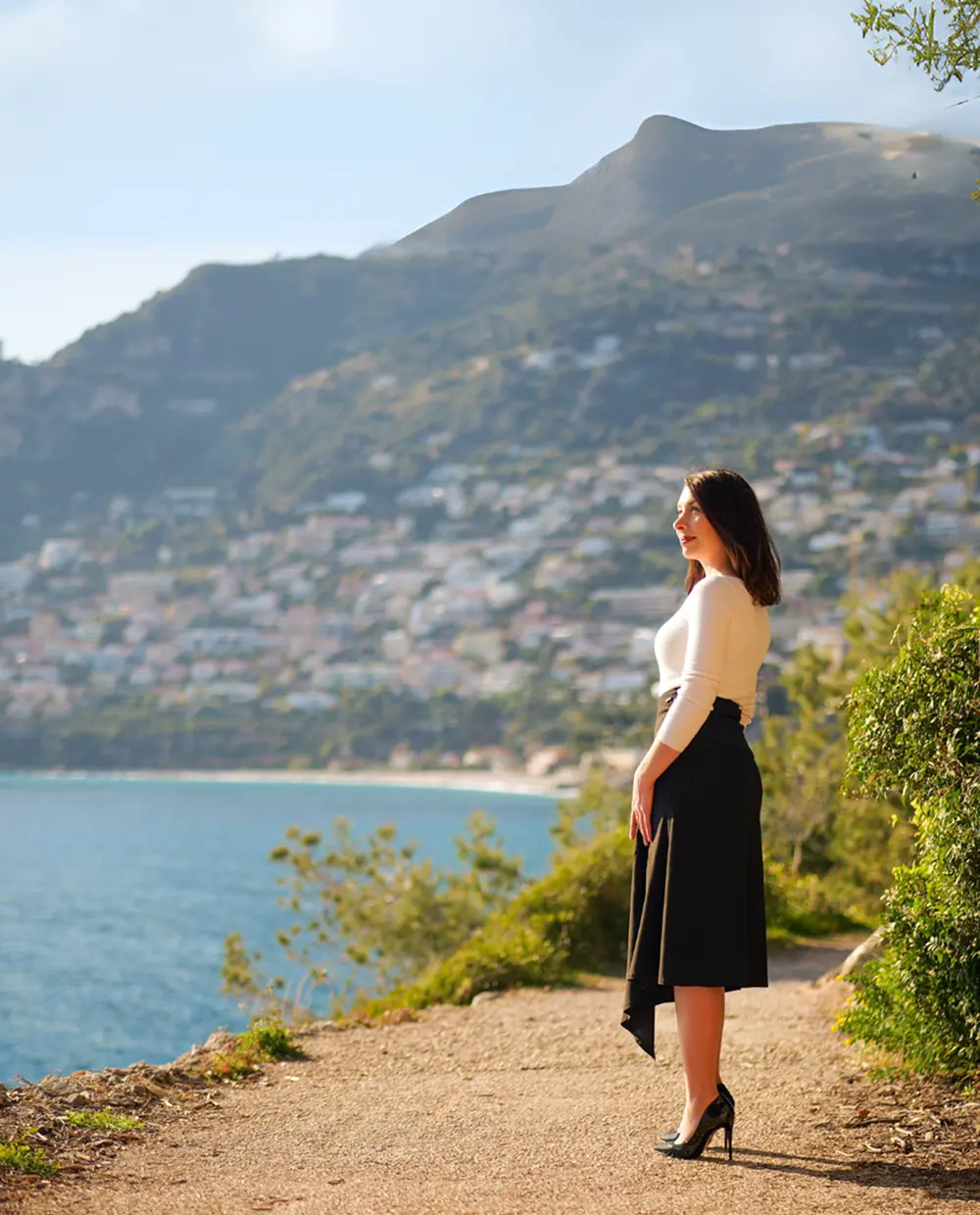 Woman in a white top and black skirt standing on a coastal path looking out at the sea with hills and buildings in the background.