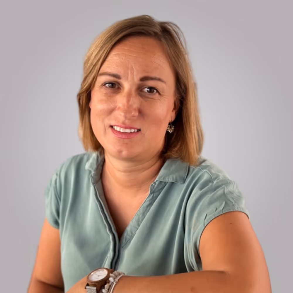Helena. Smiling woman with short blonde hair wearing a green top and a watch on her left wrist.
