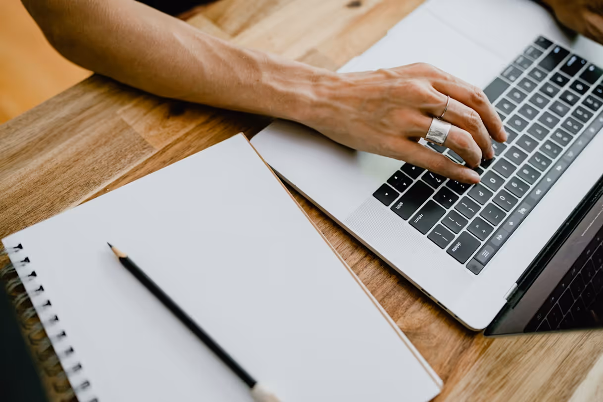 Person typing on a laptop keyboard beside a blank spiral notebook and pencil on a wooden table.