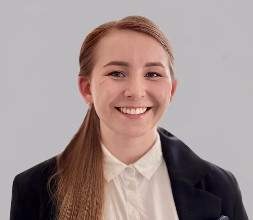 Polina. Smiling woman with long hair in a ponytail wearing a white shirt and black blazer holding a folder.