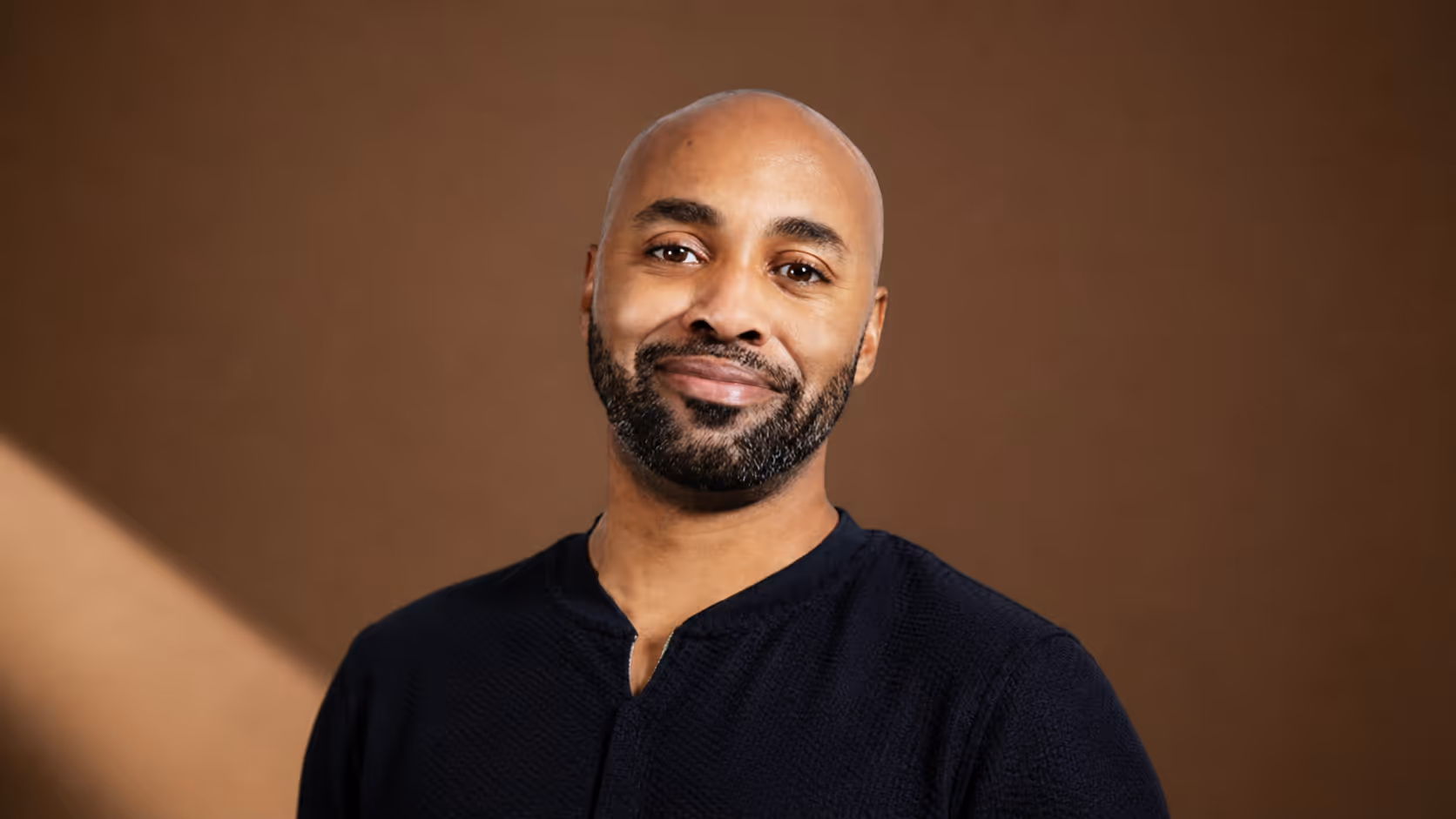 Smiling bald man with beard wearing black shirt against a brown background.