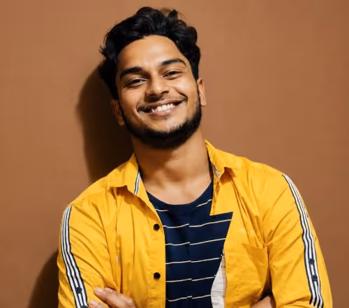Smiling young man with dark hair wearing a yellow shirt over a striped black T-shirt against a brown background.