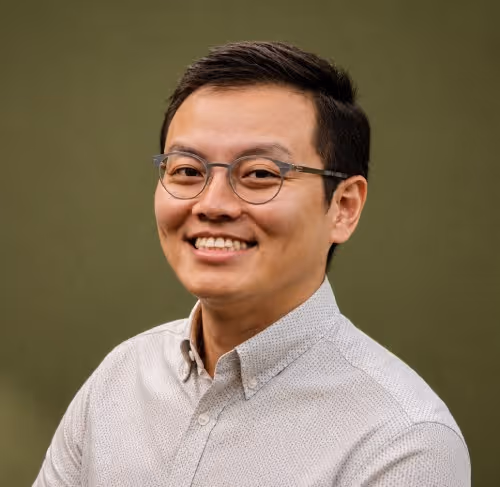 Smiling Asian man wearing glasses and a light gray button-up shirt against a green background.