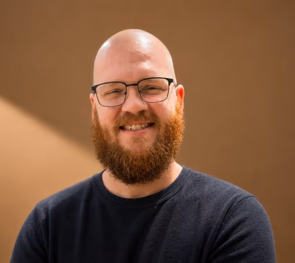 Smiling bald man with glasses and a full beard wearing a dark shirt against a plain brown background.