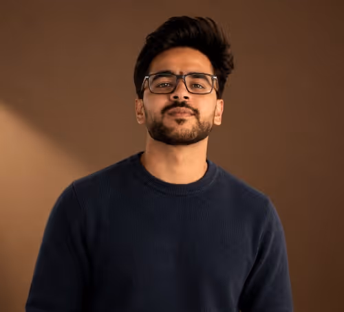 Young man with glasses and beard wearing a navy blue sweater against brown background.