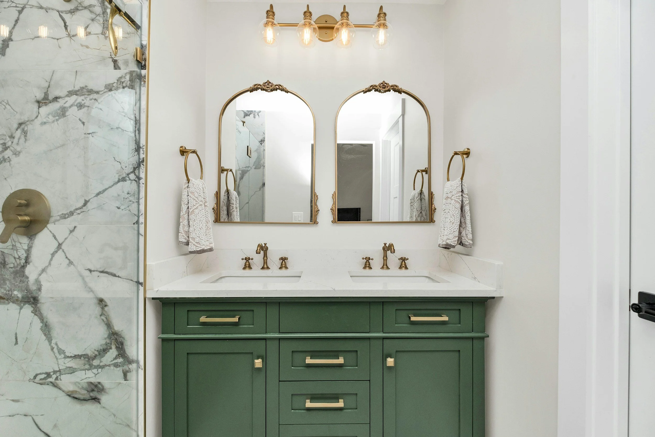 Bathroom with green double vanity, two gold-framed mirrors, brass fixtures, and marble shower wall.