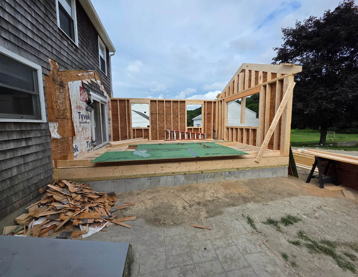 Wooden frame construction of a house extension with green floor panels and ladder leaning inside.