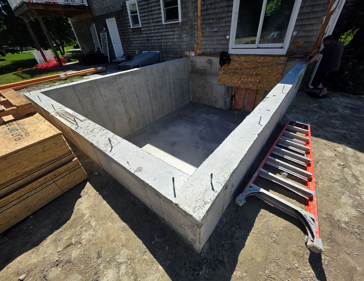 Concrete foundation pit next to a house under construction with a folded ladder nearby.
