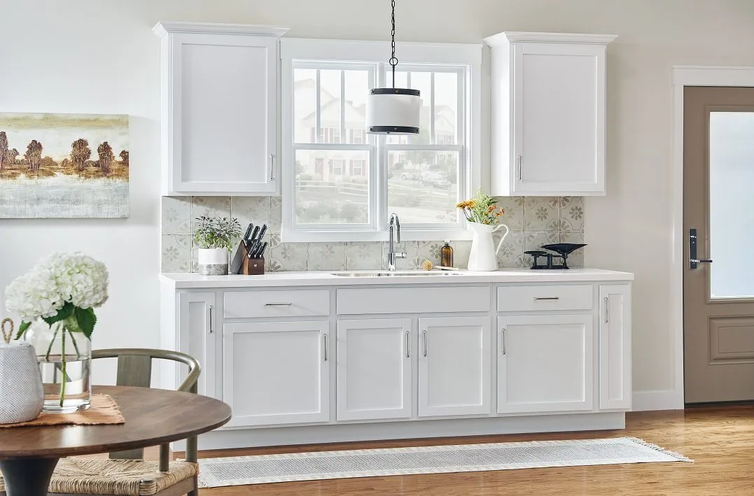 Bright kitchen with white cabinets, a sink under window, countertop decor, wooden floor, and round table with flowers.
