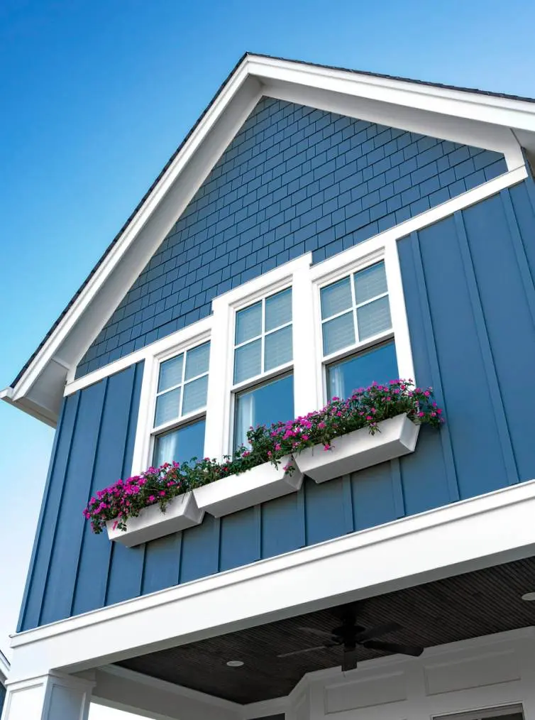 Upper blue siding of a house with white-trimmed windows and flower boxes with pink flowers.