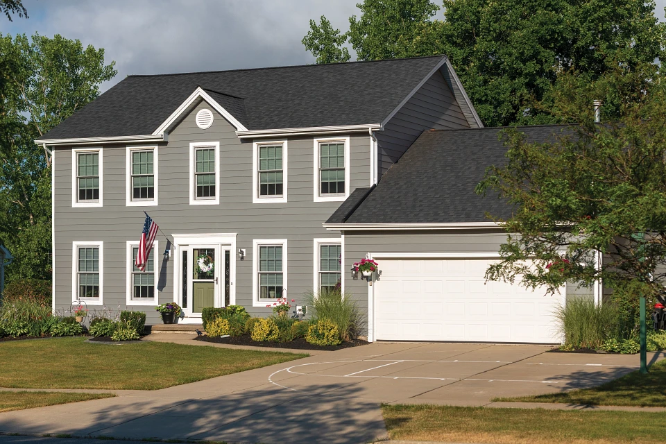 Two-story gray house with white trim, green door, American flag, and attached garage on a sunny day.