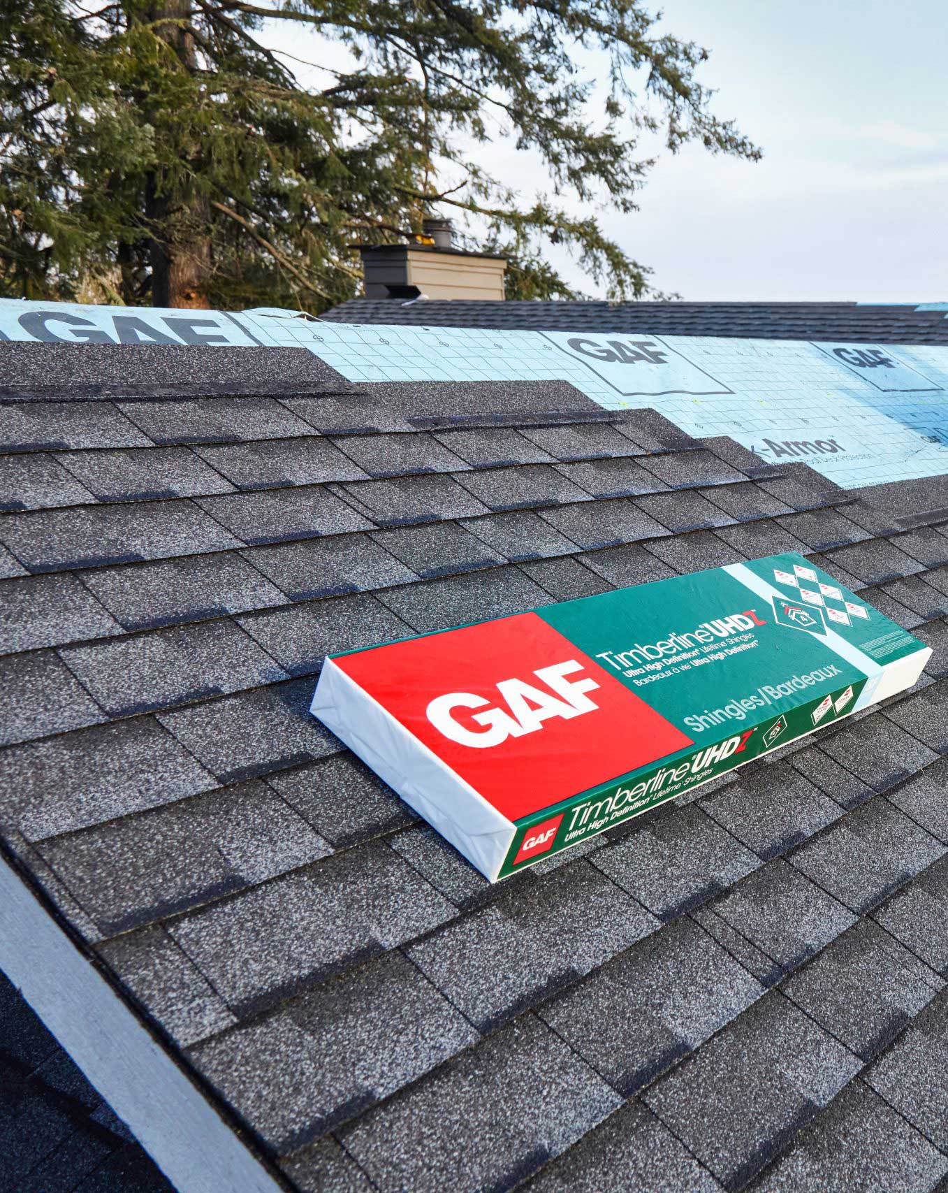 Box of GAF Timberline UHD shingles on a partially shingled roof under a clear sky.