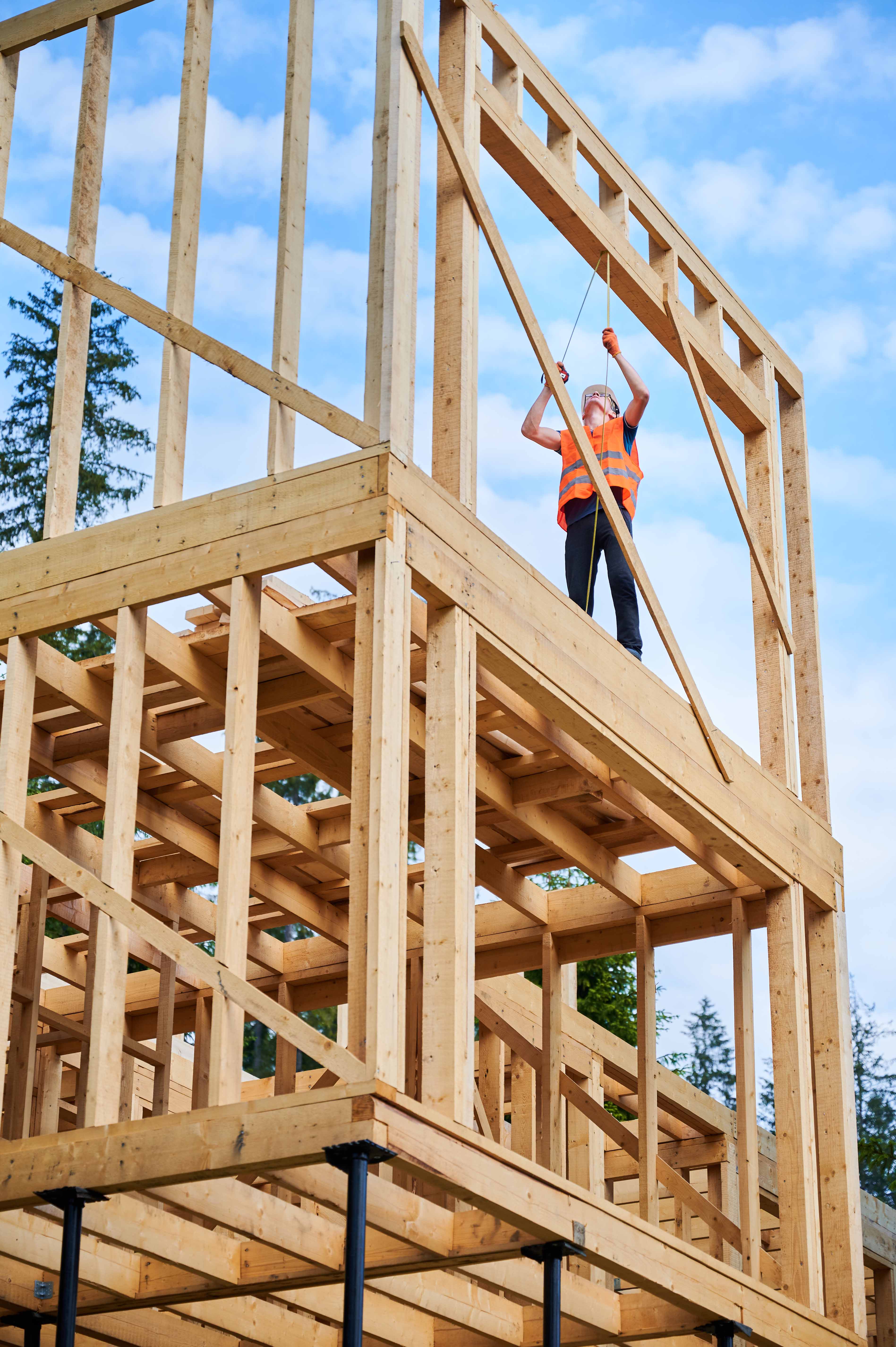 Carpenter in orange vest measuring wooden frame of building under construction against blue sky.