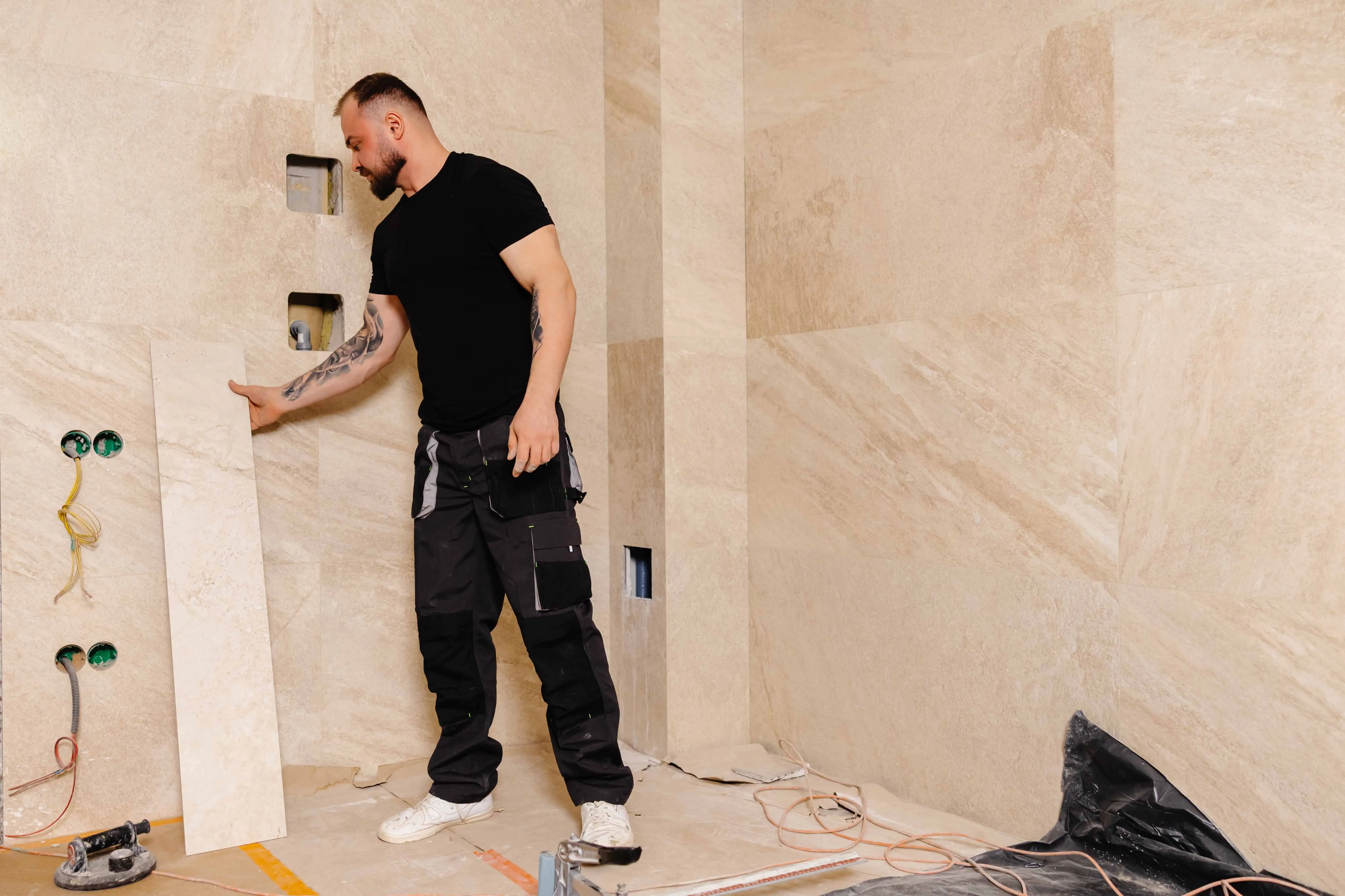 Man installing large beige wall tiles in a room under construction.