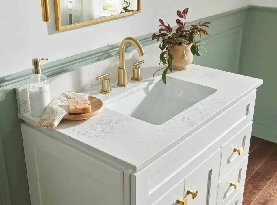 White bathroom vanity with gold faucet and handles, soap dispenser, folded cloth, and potted plant on countertop.