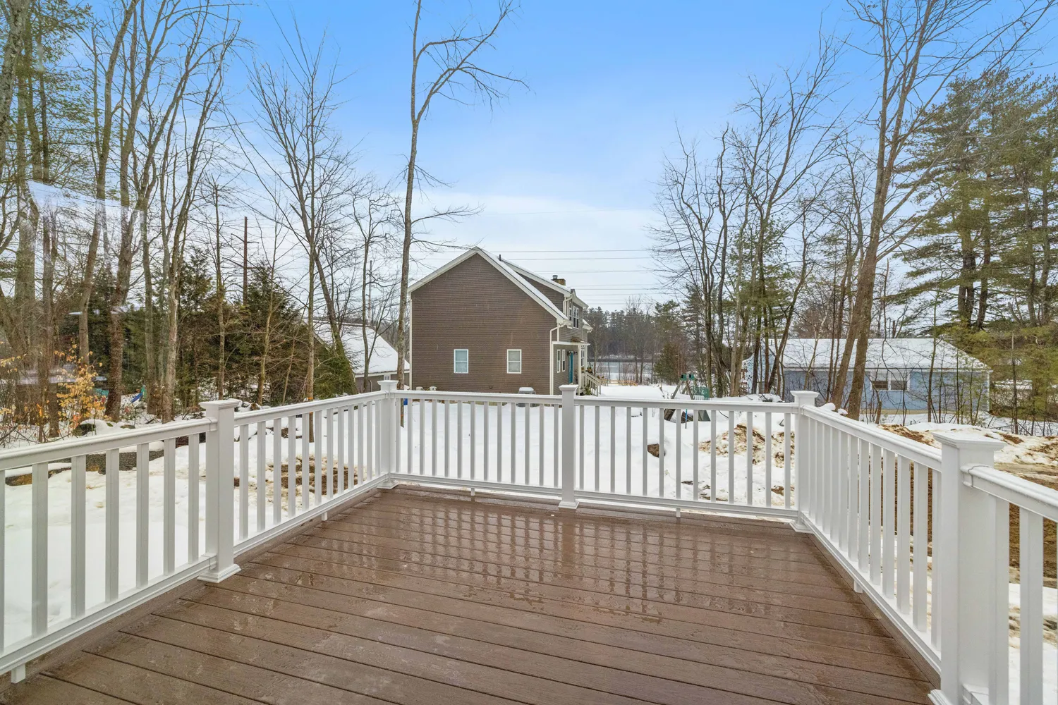 Wooden deck with white railing overlooking a snowy backyard and houses with leafless trees.