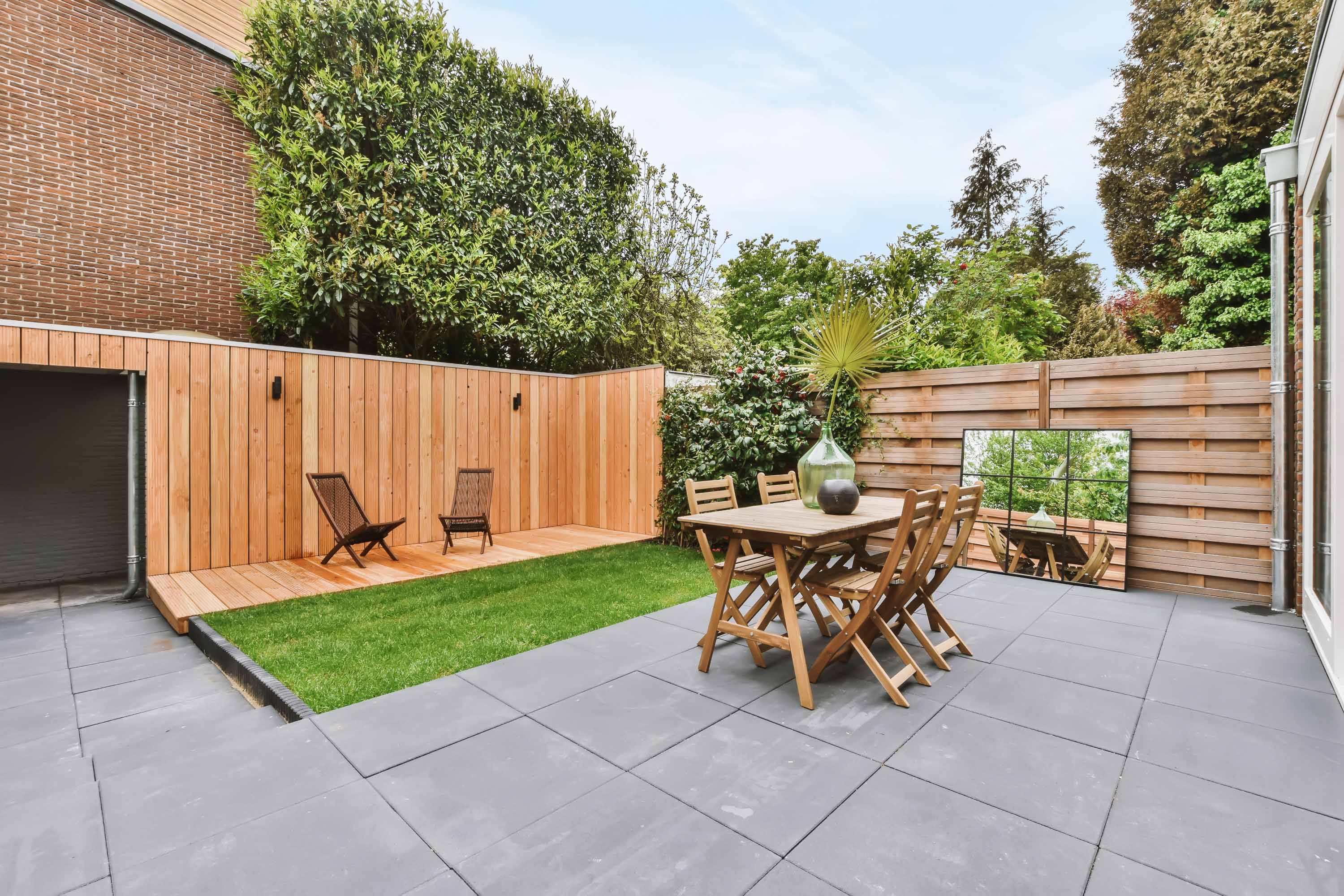 Outdoor patio with gray tiles, wooden dining set, lawn, two wicker chairs, and wooden privacy fences.
