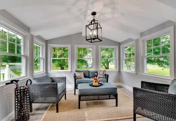 Bright sunroom with wicker furniture, gray cushions, large windows, and a rectangular coffee table.