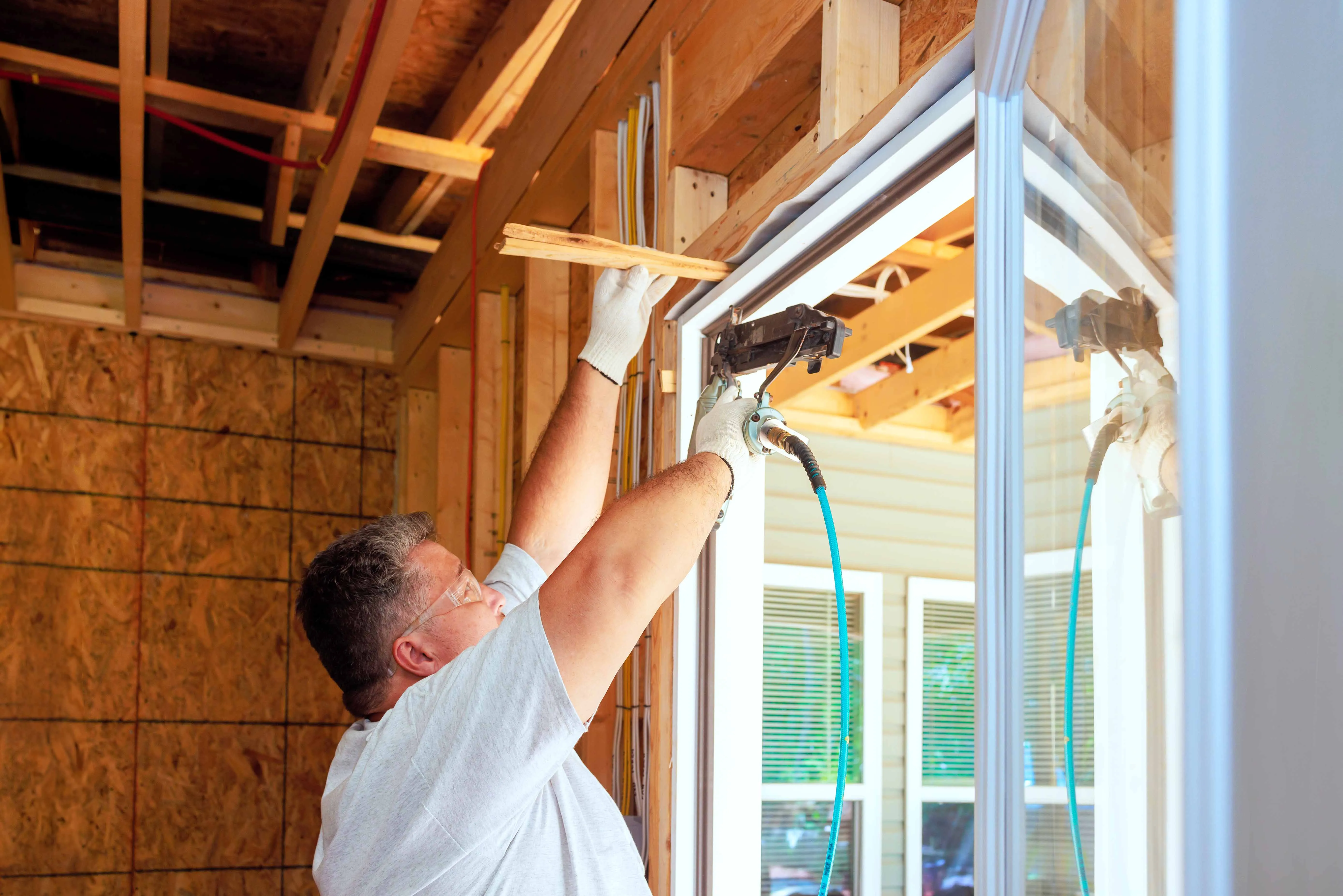 Construction worker wearing gloves and glasses installing a window frame with a nail gun.