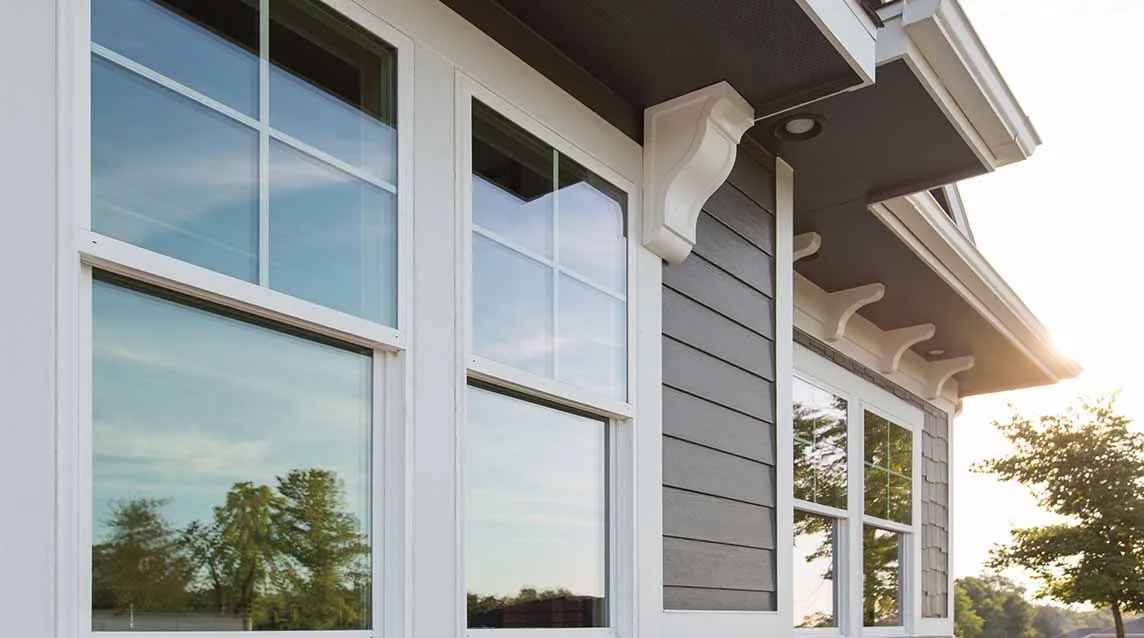 Exterior of a house showing white-trimmed double-hung windows reflecting trees and sky.
