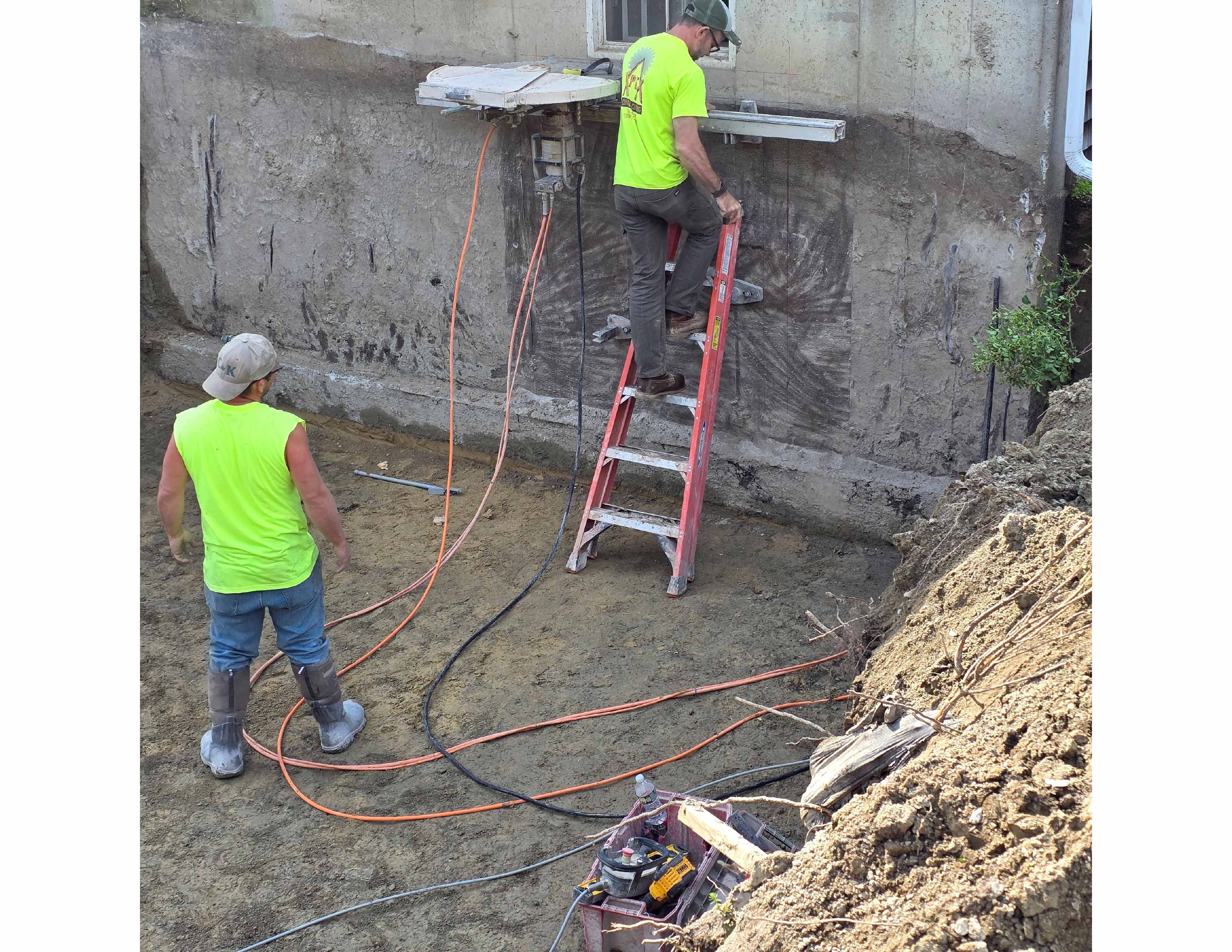 Two construction workers in neon shirts working near a concrete wall with cables and a ladder.