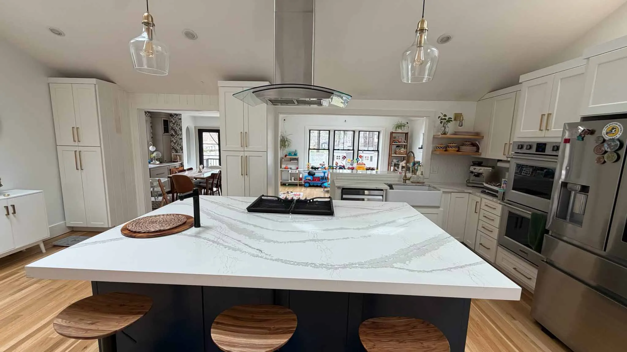 Modern kitchen with white marble island, wooden stools, stainless appliances, and large window in playroom.