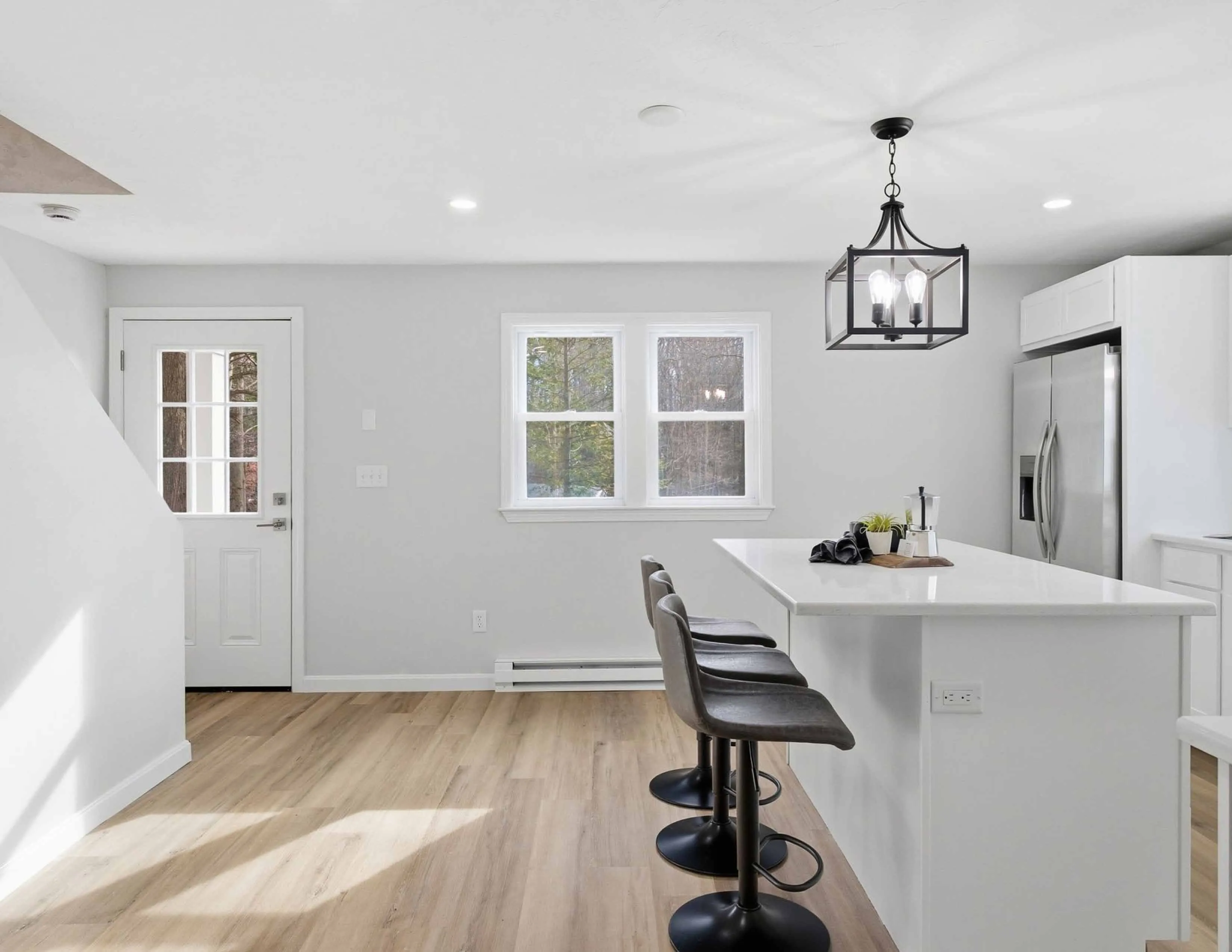 Bright modern kitchen with white island, three black bar stools, stainless refrigerator, and light wood floor.