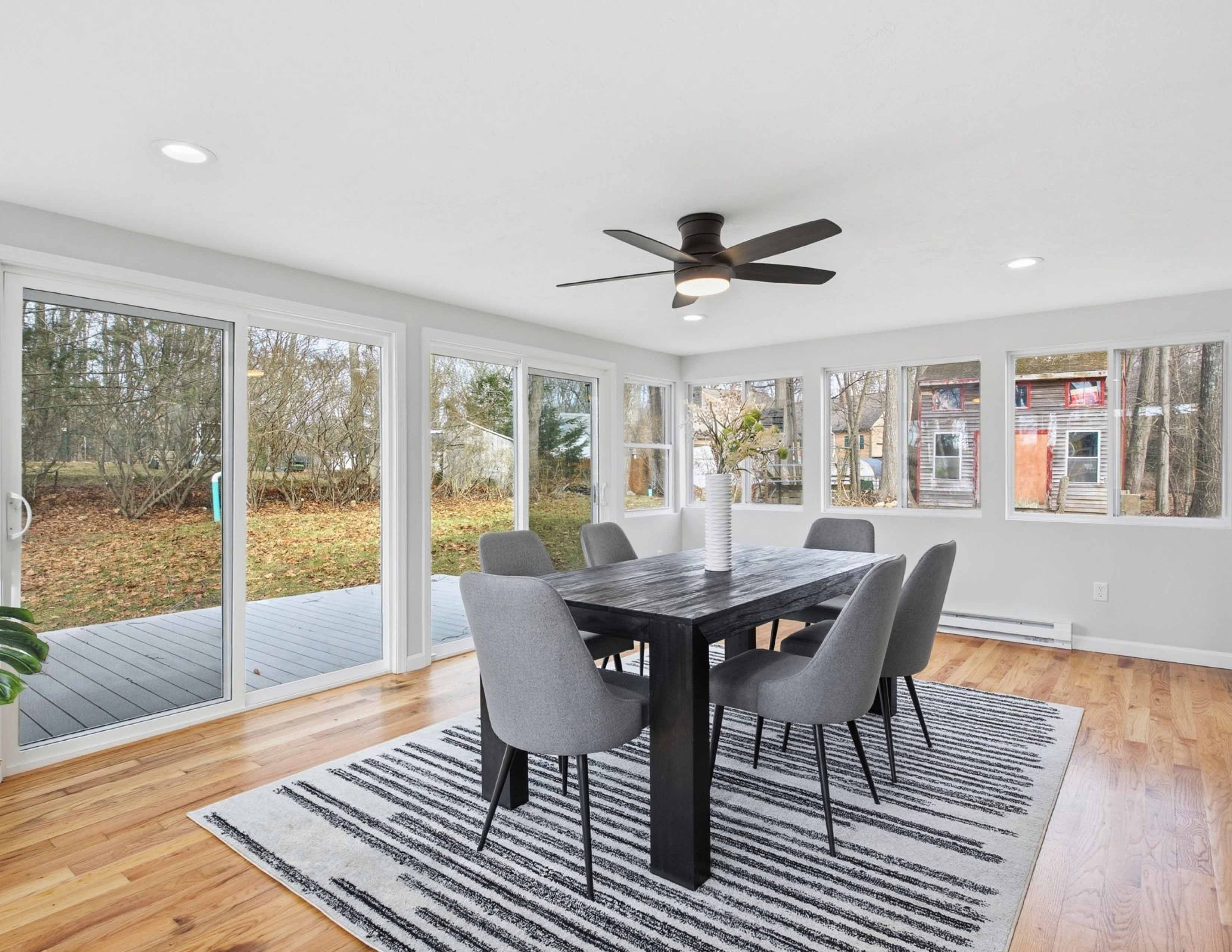 Modern dining room with black table, six gray chairs, striped rug, ceiling fan, and large windows.