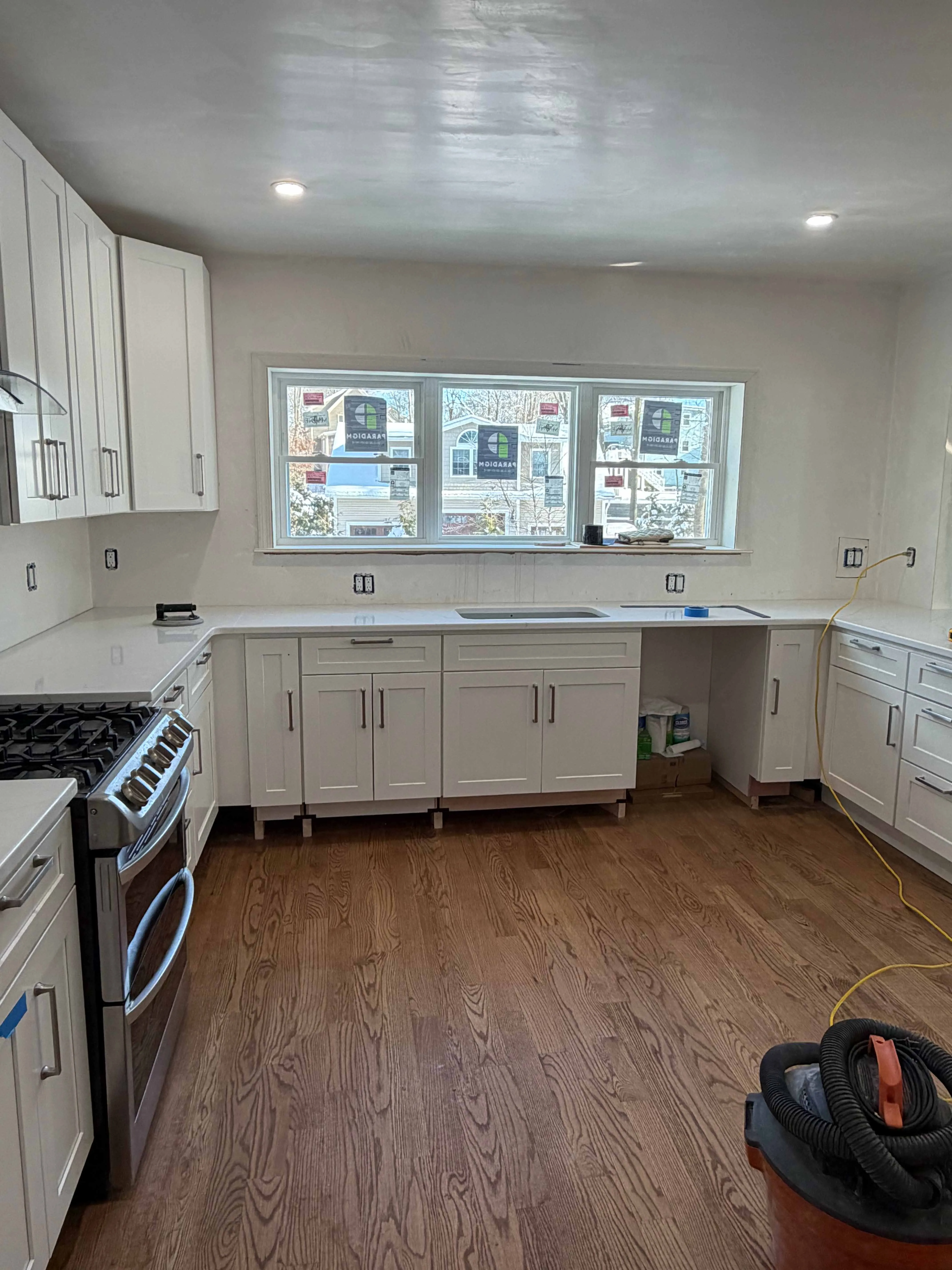 Modern kitchen under construction with white cabinets, countertop, stove, wood floor, and large window.