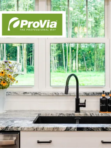 Kitchen sink with black faucet and granite countertop below white window overlooking green trees.