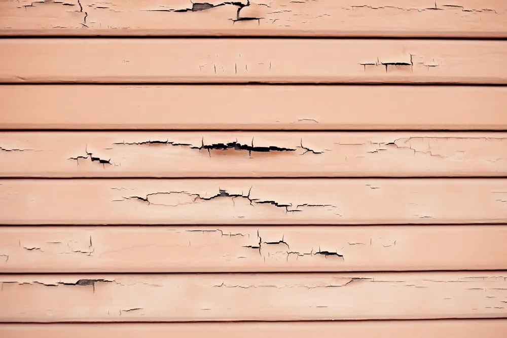 Peach-colored wooden siding with peeling and cracked paint on horizontal panels.
