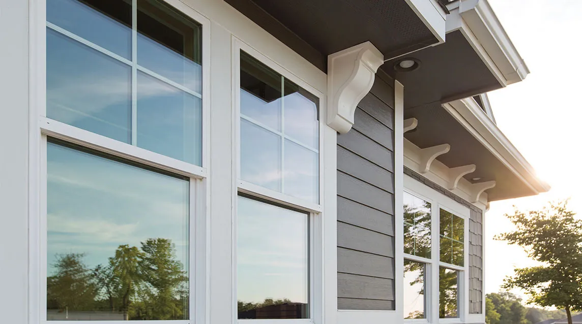 Close-up of white framed windows on a gray house exterior reflecting trees and sky at sunset.