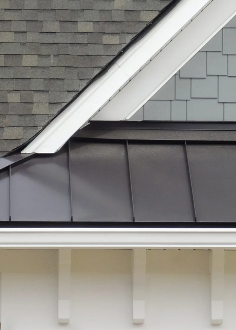 Close-up of house roof with a modern mix of black metal roofing panels, gray shingles, and white trim details.