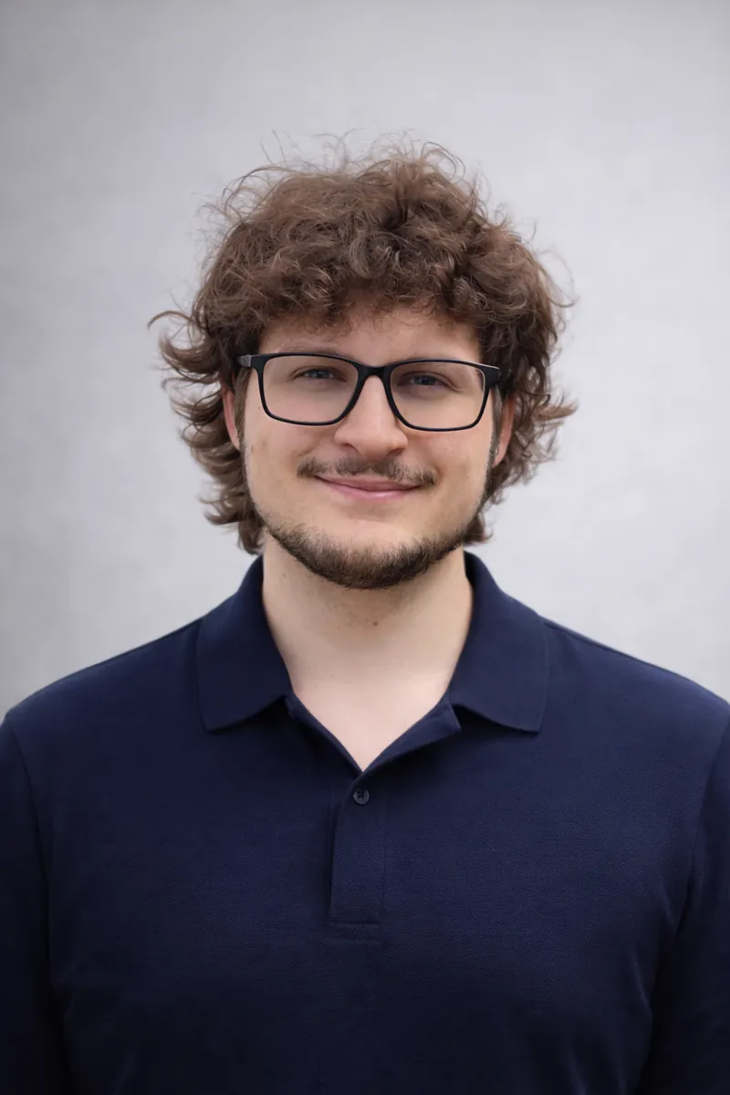 Young man with curly brown hair, glasses, and a beard smiling, wearing a navy blue polo shirt against a plain light background.
