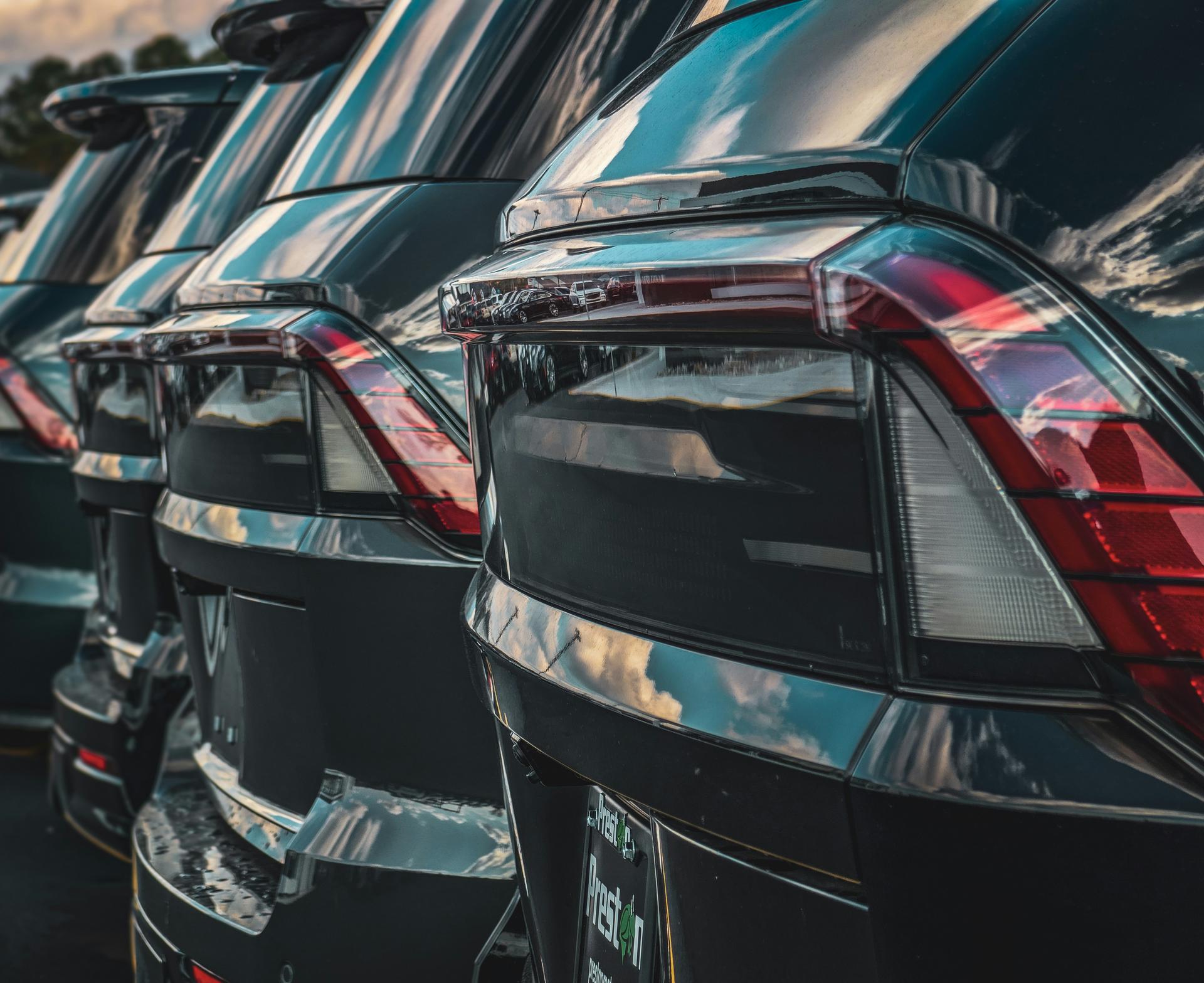 Close-up view of the rear ends of multiple black cars parked in a row with reflections of the sky on their surfaces.